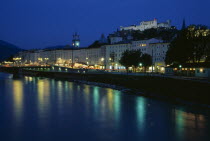 City view at night with green and yellow lights of river side street lamps and buildings reflected on water surface.  Hohensalzburg Fortress elevated on hillside behind.