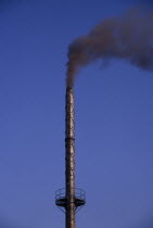 Tall factory chimney emitting black smoke against blue sky.