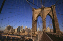 Brooklyn Bridge.  View across bridge  pedestrians and cyclist to Manhattan skyline part framed by stone tower and intersected by steel wires and suspension cables.Spans the East River.  Construction...