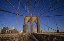 Brooklyn Bridge.  View along bridge to pedestrians at far end and Manhattan skyline beyond intersected by steel wires and suspension cables.