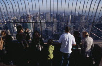 View north over city  from top of the Empire State Building at 8.30 a.m. with line of visitors looking out through expanse of glass.  One person using camcorder.