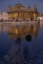 Sikh couple sitting on marble walkway looking out across sacred pool towards the Golden Temple and shimmering reflection on rippled water surface.Asia Asian Bharat Inde Indian Intiya Reflexion Religi...