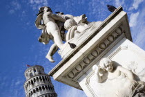 The Campo dei Miracoli or Field of Miracles.The Leaning Tower or Torre Pendente belltower with tourists on the top level under a blue sky with a statue and water fountain with cherubs in the foregroun...