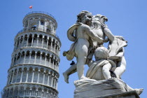 The Campo dei Miracoli or Field of Miracles. The Leaning Tower of Torre Pendente belltower with a statue of three cherubs in the foreground under a blue skyEuropean Italia Italian Southern Europe Tos...