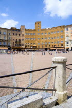 The Piazza del Campo and surrounding buildings with tourists walking in the square. Three lines in flat stone laid into the herringbone brick pattern of the square radiate from the central drain towar...