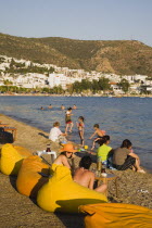 Line of brightly coloured bean-bags set out along beach for late afternoon cafe and restaurant trade.  White painted buildings spread across hillside beyond.Turkish Aegean coastmediterraneanresort...