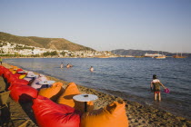 Line of brightly coloured bean-bags set out along beach for late afternoon cafe and restaurant trade.  People in shallow water along shoreline.  White painted buildings spread across hillside beyond....