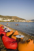 Line of brightly coloured bean-bags set out along beach for late afternoon cafe and restaurant trade.  People in shallow water along shoreline.  White painted buildings spread across hillside beyond....