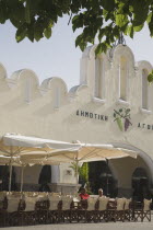 Plateia Eleftherias Kos Town Square with market entrance behind outside cafe tables beneath sun shade in foreground.Greek IslandsResort Summer Clear Blue Sky early season Aegean Dodecanese Destinati...