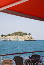 Outside cafe tables under red awning looking out across sea towards cruise boats and rocky coastline.Turkish Aegean coastOttomanMediterraneanresortearly summerseaseasonHolidayDestination Des...