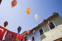 Lines of coloured lanterns and Turkish flags hanging across street from part seen  pastel painted buildings against clear blue sky.TurkishAegeancoastresortSummersunshineearly Summer seasonholi...