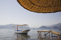 Local water taxi moored at wooden jetty from shade of part seen straw parasol in early Summer sun with calm  flat water.Turkish Rivieracoastcoastalresortsunshinesun umbrellaholidaydestination...