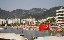 Turkish flag flying from roof of water taxi with tourists and resort beach and apartments behind in early Summer sun.Turkish Rivieracoastcoastalresortsunshinesun umbrellaholidaydestinationwat...