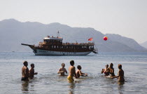 Family of Turkish tourists playing with beach ball in calm water with day cruise boat behind in late afternoon sun in early Summer season.Turkish Rivieracoastcoastalresortsunshineholidaydestina...