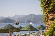 Parasols and sunbeds for hire on beach with views over bay and moored Gulet on clear bright early Summer day.  Part seen palm tree in foreground.formerly Greek Physkos in CariaTurkish Rivieracoast...