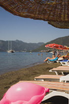 Parasols and striped sunbeds for hire on beach with views over bay on clear bright early summer day.  Tourists at waters edge with bright pink coloured beach toy and part seen sun umbrella in foregrou...