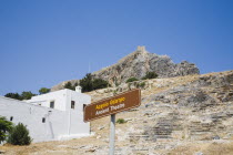 Lindos.  Popular resort village on the south of Rhodes Island.  View towards fortified hilltop castle from Ancient Theatre area with traditional Greek white-washed holiday cottage.  Sign for Ancient T...