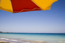 Ixia beach resort popular for water sports with part seen red and yellow striped sun umbrella in foreground against backdrop of cloudless blue sky and distant Turkish coast on bright summer morning....