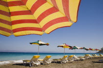 Ixia beach resort popular for water sports with red and yellow striped sun umbrella in foreground against blue  cloudless sky.  Line of sun loungers and parasols along beach beyond.AegeanRodiseaco...