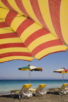 Ixia beach resort popular for water sports with red and yellow striped sun umbrella in foreground against blue  cloudless sky.  Line of sun loungers and parasols along beach beyond.AegeanRodiseaco...