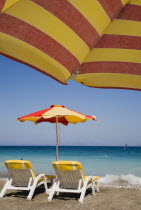 Ixia beach resort popular for water sports with red and yellow striped sun umbrella in foreground against blue  cloudless sky.  Sun loungers and parasol overlooking sea beyond.AegeanRodiseacoast c...