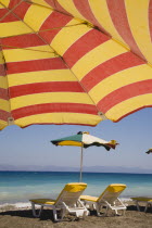 Ixia beach resort popular for water sports with red and yellow striped sun umbrella in foreground against blue  cloudless sky.  Sun loungers and parasol overlooking sea beyond.AegeanRodiseacoast c...