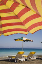 Ixia beach resort popular for water sports with red and yellow striped sun umbrella in foreground against blue  cloudless sky.  Sun loungers and parasol overlooking sea beyond.AegeanRodiseacoast c...