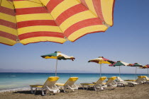 Ixia beach resort popular for water sports with part seen red and yellow striped sun umbrella in foreground against blue  cloudless sky.  Line of sun loungers and parasols overlooking sea beyond.Aege...