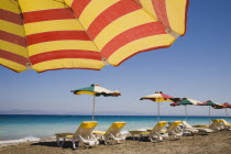 Ixia beach resort popular for water sports with part seen red and yellow striped sun umbrella in foreground against blue  cloudless sky.  Line of sun loungers and parasols overlooking sea beyond.Aege...