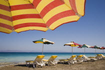 Ixia beach resort popular for water sports with part seen red and yellow striped sun umbrella in foreground against blue  cloudless sky.  Line of sun loungers and parasols overlooking sea beyond.Aege...