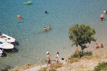 Lindos.  View over clear shallow water of bay with moored boats and tourists swiming in late afternoon sunshine.  Path to beach  rocky ground and small tree in foreground.Greek IslandsAegeanRodico...