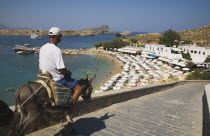 Lindos.  Donkey owner riding towards bay to bring tourists to old town and castle with lines of sun umbrellas along curve of beach beyond.Greek IslandsAegeanRodicoast coastalsearesortholidaypa...