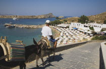 Lindos.  Donkey owner riding towards bay to bring tourists to old town and castle with lines of sun umbrellas along curve of beach beyond.Greek IslandsAegeanRodicoast coastalsearesortholidaypa...