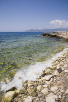 Pythagorio.  Tourists on outcrop of rock extending out into  clear water beside pebble beach with South West coast of island in distance behind.AegeanGreek IslandsPythagorionSummerseacoast coast...