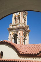 St Michael Panormitis monastery in Southern Symi.  Site of pilgrimage from the 12th Century.  Highly decorated Baroque bell tower and roof detail part seen against bright summer blue sky framed by arc...