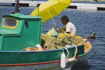 Painted fishing boat  moored in shallow water off St Michael Panormitis monastery with Greek fisherman mending nets under shade of yellow umbrella.AegeanGreek IslandsSimicoast coastalseaSummerp...