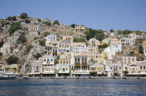 Main harbour of Yialos overlooked by pastel painted houses on waterfront and on rocky hillside behind. AegeanGreek IslandsSimicoast coastalseaSummerpackageholidayresortvacationtripdestinat...