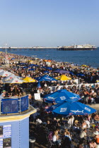 England, East Sussex, Brighton, People sitting under sun shade umbrellas at tables on the promenade outside bars and restaurants with Brighton Pier and people on the shingle pebble beach beyond.