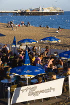 England, East Sussex, Brighton, People sitting under sun shade umbrellas at tables on the promenade outside The Beach Bar with Brighton Pier and people on the shingle pebble beach beyond.