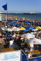 England, East Sussex, Brighton, People sitting under sun shade umbrellas at tables on the promenade outside bars and restaurants with Brighton Pier and people on the shingle pebble beach beyond.