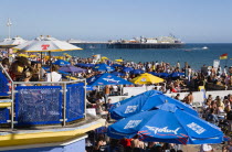 England, East Sussex, Brighton, People sitting under sun shade umbrellas at tables on the promenade outside bars and restaurants with Brighton Pier and people on the shingle pebble beach beyond.