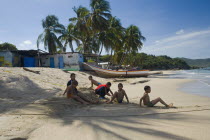 VENEZUELA, Margarita Island, Playa la Galera, Kids playing on the beach, under the shade of a palm tree just in front of the seawater while other trees, a house and a boat are noticeable at the backgr...