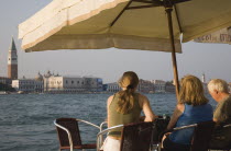 Italy, Veneto, Venice, Giudecca island, Tourists enjoying lunch at terrace cafe in Centro Storico with view to St. Marks from shade of sun umbrella in late summer.