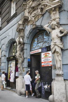 Hungary, Pest County, Budapest, Art Nouveau sculpture on facade of apartment building with local people standing in doorway below and outside shopfront on left.