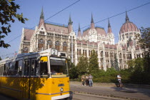 Hungary, Pest County, Budapest, yellow tram passing the Neo-Gothic exterior facade of the Parliament Building with pedestrians on pavement beyond tram line.