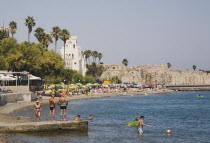 Greece, Dodecanese, Kos, Kos Town beach with castle walls and harbour entrance behind and Greek holidaymakers bathing on shingle beach in foreground.