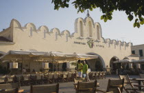 Greece, Dodecanese, Kos, Kos Town, Agora market in Summer sun with holidaymakers walking past near empty cafe seating.