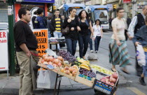 Turkey, Istanbul, Sultanahmet, Pedestrians pass fresh fruit stall with male vendor on busy street beside tramway.