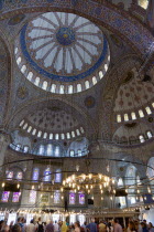 Turkey, Istanbul, Sultanahmet Camii, The Blue Mosque interior with sightseeing tourists by a chandelier below the decorated domes with stained glass windows beyond.