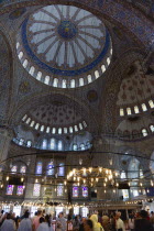 Turkey, Istanbul, Sultanahmet Camii, The Blue Mosque interior with sightseeing tourists by a chandelier below the decorated domes with stained glass windows beyond.